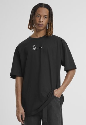 Jong persoon met middellange dreadlocks die een loszittend zwart T-shirt met de tekst "Kane" en zwarte broek draagt, hand in de zak, neutrale achtergrond.