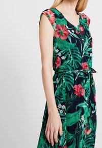 Nainen, joka on pukeutunut ilman hihuksia olevaan trooppiseen kuvioon, jossa on vihreitä lehtiä ja vaaleanpunaisia hibiskuksia, vyötäröllä solmittu vyö, seisoo yksinkertaista taustaa vasten.