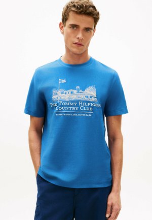 Młody mężczyzna w niebieskiej koszulce z białym nadrukiem i napisem "The Tommy Hilfiger Country Club, Sunset Ridge Lane, Silver Lake."
