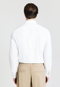 Camicia bianca a maniche lunghe con colletto classico, tessuto morbido e polsini con bottoni, abbinata a pantaloni beige con vita elastica e tasche posteriori.