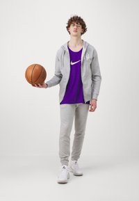Sivi hoodi, ljubičasta Nike majica bez rukava, sivi trenirka i bijele atletske tenisice. Drži košarkašku loptu, stojeći na neutralnoj pozadini.