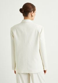 Blazer blanco a rayas con un diseño estructurado, que presenta mangas largas y un corte recto. La tela parece suave y bien ajustada.