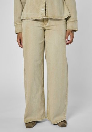 Beige Jeans mit weitem Bein, kombiniert mit einer passenden, geknöpften Jacke, die Hände entspannt seitlich, dazu sind braune Schuhe mit quadratischer Spitze zu sehen.