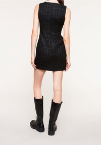 Mini abito nero in tessuto texturizzato con silhouette aderente, che presenta un colletto a giro e senza maniche. Abbinato a stivali neri fino al ginocchio.