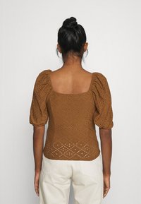 Brun, texturerad topp med puffärmar och fyrkantig halsringning, med ögla detalj och en figurnära silhuett. Bärs med ljusa byxor.