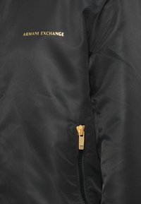 Sort jakke med en glat tekstur, guldfarvet lynlås og logo. Bemærkelsesværdige detaljer inkluderer et slankt design og et nærbillede af stoffet.