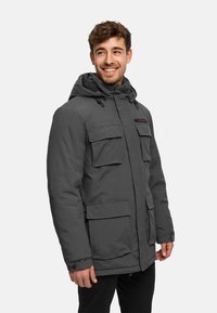 Homme souriant, portant une veste d'hiver à capuche gris foncé avec plusieurs poches frontales, debout devant un fond clair uni.