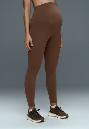 Personne enceinte portant des leggings de maternité marron et des baskets marron, debout sur un fond gris uni.