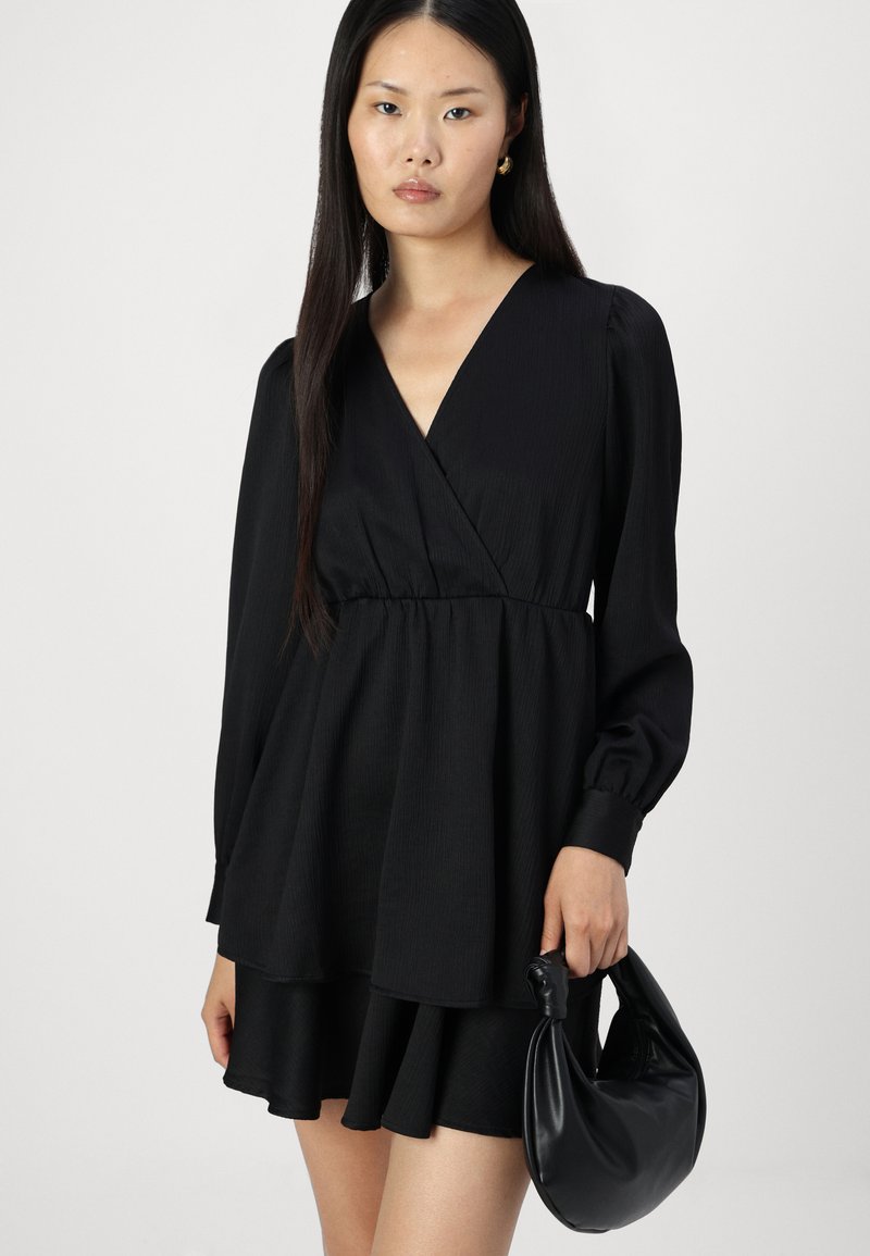 Robe noire à manches longues avec un design plissé. Associée à un sac à main noir à texture lisse. Silhouette simple et élégante.