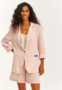 Světle růžový lněný blazer s ohrnutými rukávy, jedním knoflíkem a předními kapsami. Spárováno s odpovídajícími růžovými lněnými šortkami.