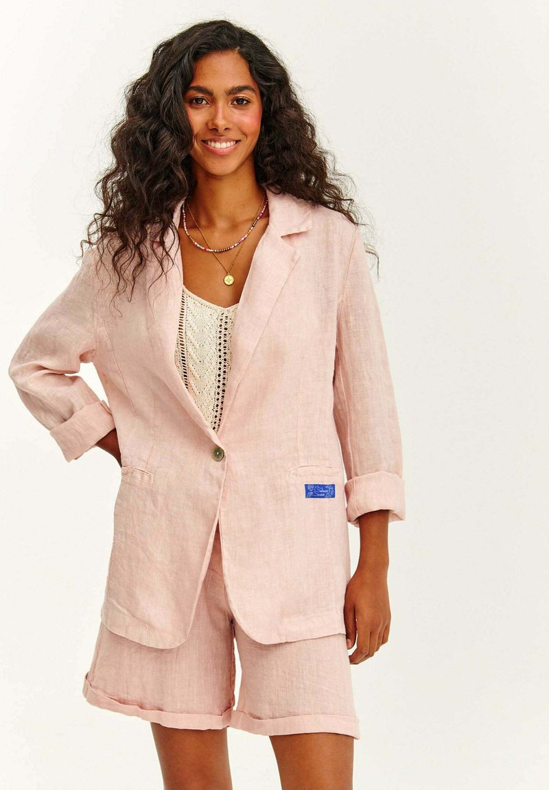 Světle růžový lněný blazer s ohrnutými rukávy, jedním knoflíkem a předními kapsami. Spárováno s odpovídajícími růžovými lněnými šortkami.