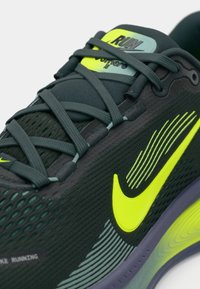Tamsiai žalios sportinės bateliai su geltonomis akcentais, tinklinis viršus, tekstūruota paviršius, išryškintas šoninis swoosh ir atitinkamos raišteliai.
