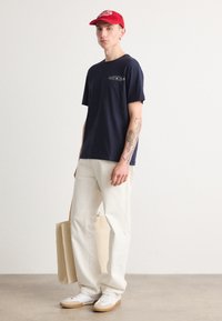 Marineblaues T-Shirt mit Drucklogo, weiße weit geschnittene Hose, rotes Cap, weiße Sneaker mit beigen Sohlen und eine beige Tragetasche.