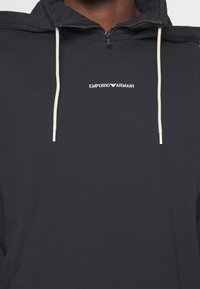 Marinblå zip-up hoodie i texturerad tyg, med en vit dragkedja och en liten vit "EMPORIO ARMANI"-logotyp på bröstet.