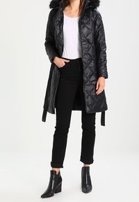 Svart, quiltad puffjacka med avtagbar huva klädd med päls, långa ärmar och knälång design, kombinerad med svarta jeans och ankelboots.