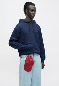 Μπλε σκούφη φούτερ με μπροστινή τσέπη και κορδόνι, με μικρό λογότυπο. Ανοιχτό μπλε τζιν παντελόνι με φαρδιά μπατζάκια και τσαλακωμένο κόκκινο καπέλο.