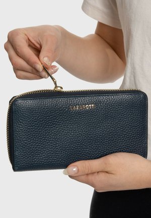 Cartera de cuero azul marino con acabado texturizado, cierre de cremallera dorada y logo de la marca. Sostenida en la mano, mostrando su forma rectangular.