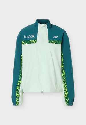 Lyseblå og blågrønn glidelåsjakke med grønne, mønstrede striper på ermer og bryst, med TCS- og New Balance-logoer.