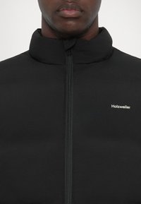 Musta puhvoroikku korkean kauluksen kanssa, etuvetoketju ja pieni valkoinen "Holzweiler" -logo vasemmassa rinnassa. Sileä, matta pintatextuuri.