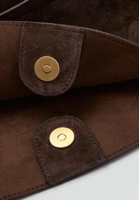 Solapa de ante marrón con dos cierres de botón dorados, que presenta una textura suave y bordes redondeados, sobre un fondo más claro.