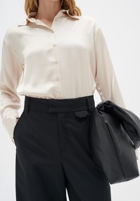 Cremfarbende Satin-Hemd mit Knopfleiste und spitzen Kragen, kombiniert mit hoch geschnittenen schwarzen Hosen mit flachem Vorderteil und Seitentaschen. Schwarze Tasche über dem Arm getragen.