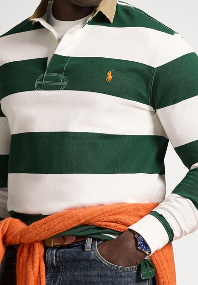 Muž v zeleno-bielom pruhovanom rugby tričku, s oranžovým svetríkom zviazaným okolo pása, modrými hodinkami a modrými džínsami s rukou v vrecku.