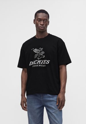 Άνδρας που φοράει μαύρο t-shirt Dickies Workwear με καρτούν χαρακτήρα και ανοιχτό μπλε τζιν, στέκεται μπροστά σε απλό ανοιχτό φόντο.