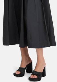 Schwarzer, plissierter Rock mit glatter Textur, kombiniert mit schwarzen, offenen Blockabsatz-Mules. Beide haben ein minimalistisches Design und klare Linien.