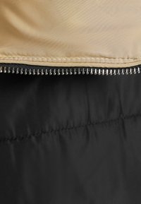 Padded jacket featuring a beige outer layer with a smooth finish and a black interior, detailed with a metal zipper and contrasting stitching.