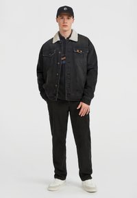 Ung man som står upprätt, klädd i en svart keps, svart denimjacka med sherpa-krage, svarta byxor och vita sneakers mot en enfärgad ljus bakgrund.
