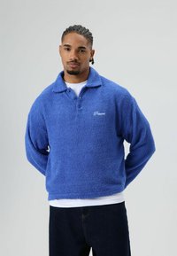 Blauwe fleece trui met kraag, lange mouwen en geborduurd merkembleem. Zachte textuur en ontspannen pasvorm, gedragen over een wit overhemd.