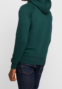 Sudadera verde con bolsillo tipo canguro y capucha ajustable con cordón, combinada con jeans de mezclilla oscura. Puños y dobladillo acanalados.