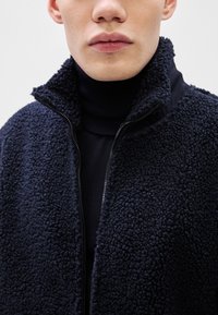 Tumesinist fliisjakk kõrge kraega, tekstuurse pinnaga ja täislukuga. Kantud musta polokaelusega üle, rõhutades värvide kontrasti.