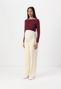 Graviditetsoutfit med en vinröd långärmad tröja och krämfärgade vida byxor med justerbart elastiskt midjeband och dragsko.