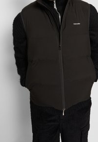 Schwarze Puffer-Weste mit hohem Kragen und Reißverschluss vorne. Glatte Stofftextur mit Steppdetails. Logo "Holzweiler" auf der linken Brust.