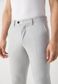 Slim-fit světle šedé kalhoty se bočními kapsami a černým knoflíkem, které má na sobě osoba ve bílé košili zastrčené do kalhot.