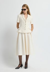 Donna che indossa un blazer corto crema a maniche corte e una gonna midi plissettata abbinata, collant neri, scarpe con tacco T-strap nere e occhiali da sole a occhio di gatto.
