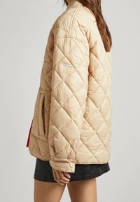 Chaqueta beige acolchada con forma cuadrada. Cuenta con un bolsillo lateral y un forro interior rojo. Textura suave con una etiqueta sencilla en la manga.