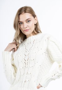 Pull en tricot blanc présentant un design en grosse maille à torsades avec des motifs ajourés, un col rond et des poignets côtelés.
