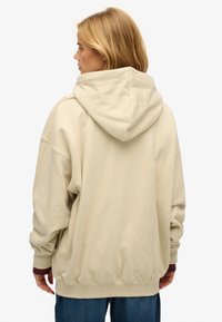 Kobieta o blond włosach, ubrana w oversize'ową beżową bluzę z kapturem i niebieskie dżinsy, zwrócona plecami, na jednolitym białym tle.