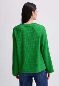 Grüner strukturierter Pullover mit Rundhalsausschnitt, Raglanärmel und lockerer Passform. Waffelmuster, kombiniert mit Jeans.