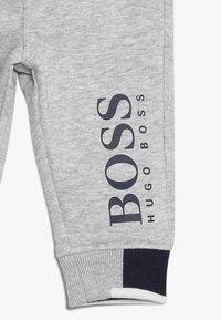 Šedé tepláky vyrobené z měkké bavlny s námořnickým logem "BOSS HUGO BOSS" tištěným na straně. Obsahují žebrované manžety a kontrastní akcenty.