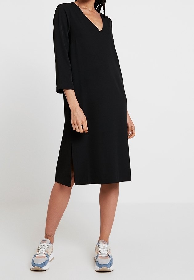 Femme portant une robe noire à col en V, longueur genou, avec manches trois-quarts et fentes latérales, associée à des baskets blanches et bleues.