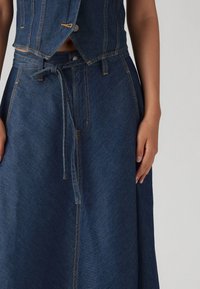 Džínová crop top a dlouhá sukně v tmavě modré barvě. Vlastnosti zahrnují viditelné švy, zavazovací opasek a přiléhavý pas. Hladká textura.