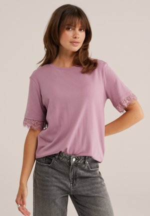Femme portant un t-shirt lavande à manches courtes avec bordure en dentelle aux manches et un jean noir délavé taille haute, posant avec une main sur la hanche.