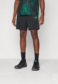 Black mesh athletic shorts with a green Adidas logo, paired with black shoes and black crew socks. The fabric has a textured finish.