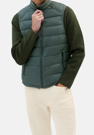 Mężczyzna w ciemnozielonej pikowanej kamizelce bez rękawów, nałożonej na oliwkową, długorękawową koszulę, beżowe spodnie, jedną ręką w kieszeni, drugą dotykający kołnierza.