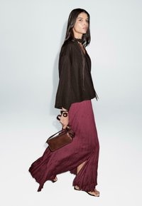 Haut marron ample avec des liens, longue jupe plissée bordeaux, sac en cuir marron à la main et chaussures noires minimalistes.