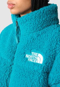 Tyrkysová fleecová bunda s vysokým límcem, zapínáním na zip a bílým vyšívaným logem. Měkká textura a pohodlný design.