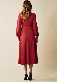 Lang, rød glitrende kjole med en tætsiddende overdel, puffede ærmer og en udvidet nederdel. Sorte højhælede sko fuldender outfittet.